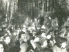 2nd Jewish Song Festival, Succot, 1979. More participants attended this time. There are a lot of young people in the audience. The symbolic sukkah served as a hall for the festival. Pine tree branches hang on the ropes. Policemen are standing behind the branches (they can be seen in the background on the left side). Refuseniks are scattered throughout the audience: at the wall of the sukkah (in the center)  Sasha Kremen (boy), next to him are his mother, Galya Kremen, and Galya Gurevich. In the second row (to the left), sitting and attentively looking at the stage is Kira Razgon a singer and attendee of all the festivals. In the first row (to the right), in profile (without a hat) is Lena Gurevich. Moscow, Ovrazhki forest, Succot, 1979. co RS